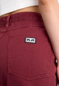 Jeans din denim maroniu care prezintă o buzunar din spate cu o etichetă logo alb-negru, cusături vizibile și un material cu textură netedă.