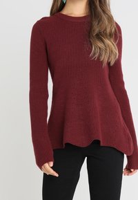 Donna che indossa un maglione a coste bordeaux con maniche lunghe e un orlo leggermente svasato, abbinato a pantaloni neri.