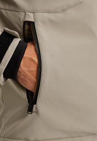 Beige bunda s kapsou na zip. Kapsa má černé podšívky a zlatý zip. Vnitřní rukáv ukazuje kontrast s černým materiálem.