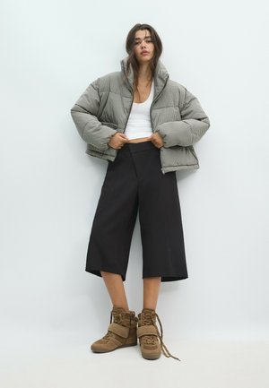 Femme portant une veste matelassée courte à carreaux, un débardeur blanc, un large pantalon culotte noir et des baskets montantes beige avec des lacets non noués.
