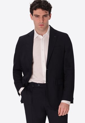 Uomo in abito nero e camicia bianca con bottoni, in piedi con una mano in tasca, guarda direttamente davanti a uno sfondo semplice.