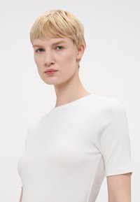 Top blanco de manga corta en tejido suave con cuello redondo. Textura lisa y forma ajustada sin costuras ni patrones visibles.