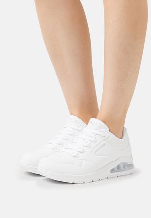 Persona che indossa sneakers bianche Skechers con suole ammortizzate su uno sfondo neutro, mostrando le gambe inferiori e i piedi.