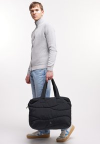 Svart quiltad väska med dubbla handtag, med dragkedjestängning och strukturerad design. Modellen bär en grå tröja och ljusa jeans.