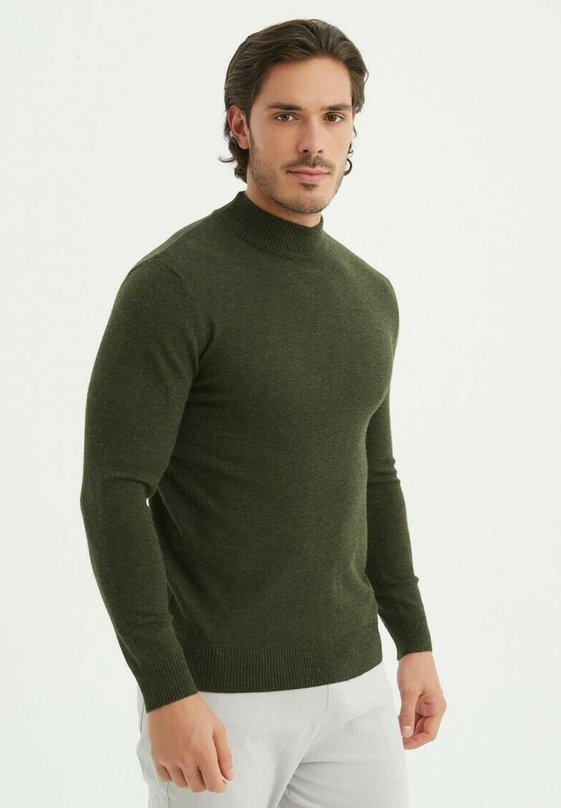 Pull en tricot vert foncé avec un col haut, une coupe ajustée et des poignets et un ourlet côtelés. Texture lisse, matériau léger et forme épurée.