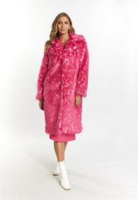 Cappotto in pelliccia sintetica rosa con un motivo testurizzato, colletto oversize e due bottoni. Indossato sopra un capo rosa coordinato. Stivaletti bianchi alla caviglia.