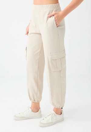 Beige cargo pants with elastic ankles worn with white sneakers, one hand in pocket, shown from waist to feet on a plain background.