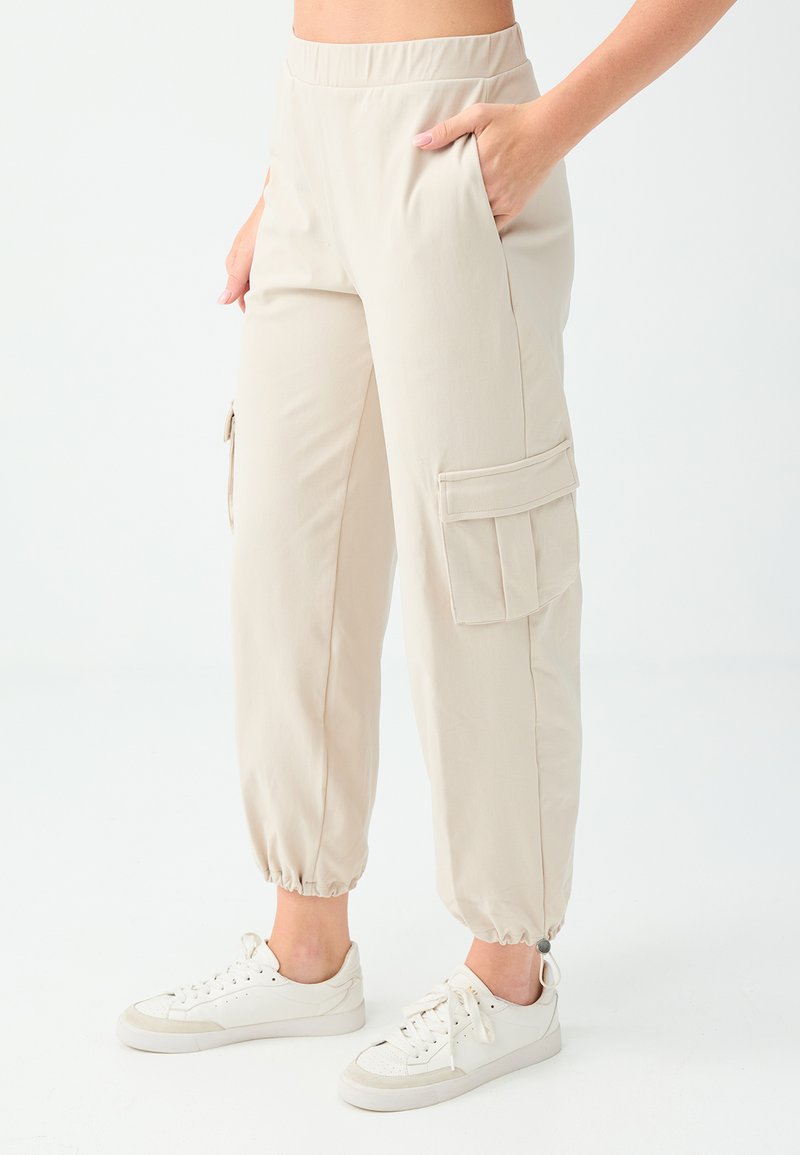 Pantalon cargo beige avec chevilles élastiques porté avec des baskets blanches, une main dans la poche, montré de la taille aux pieds sur un fond uni.