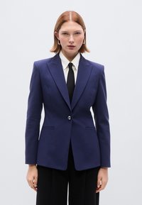 Σκούρο μπλε σακάκι με ραμμένα κομμάτια και δομημένους ώμους, με ένα ασημένιο κουμπί και δύο μπροστινές τσέπες, φορεμένο πάνω από λευκό πουκάμισο και μαύρη γραβάτα.