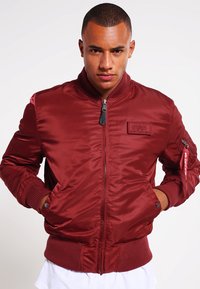 Giacca bomber in satin rosso con polsini e orlo a coste, chiusura zip frontale, tasche laterali e una toppa sul petto. Presenta un clip decorativo sulla manica.