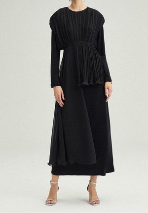 Frau trägt ein langes schwarzes plissiertes Kleid mit langen Ärmeln und silbernen offenen Sandalen, steht vor einem schlichten hellen Hintergrund.