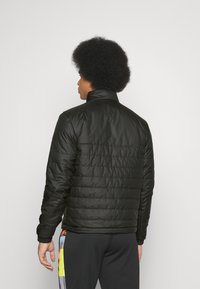 Sort polstret jakke med høj krave, med vertikal quilted tekstur og elastiske manchetter. Båret med mørke bukser, der fremviser farverige accenter.