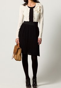 Vit stickad kofta med knappstängning och blommigt dekoren på axeln, i kombination med en svart plisserad kjol och svarta strumpbyxor. Brunt handväsk.