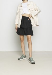 Person som bär en vit kort topp, ljusbeige denimjacka, svart lagerkjol och gröna och vita sneakers, stående mot en enkel ljus vägg.