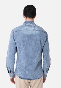 Camicia in denim azzurro chiaro con maniche lunghe, polsini a bottoni, dettagli sulla yoke posteriore e un orlo leggermente curvo. Tessuto testurizzato con un leggero effetto sbiadito.