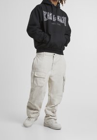 Svart hoodie med "KARL KANI EST. 1989"-logotyp, tillsammans med beige cargobyxor med sidofickor och manschetter, kompletterat med ljusa sneakers.