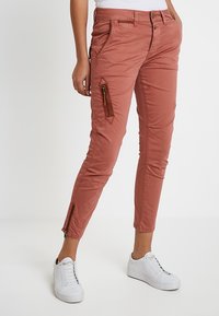 Rostfärgade slim-fit byxor med sid- och ankeldragkedjor, bärs med vita sneakers och en vit topp.