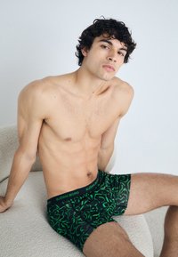Green patterned boxer shorts feature a black base, elastic waistband with "BJÖRN BORG" logo. The texture appears smooth and stretchy.