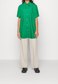 Camicia verde a maniche corte con bottoni, dotata di taschino frontale, abbinata a pantaloni beige a gamba larga e scarpe platform nere.