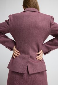 Houndstooth-mønstret blazer i burgundy og lyserøde nuancer, med klapkrave og rygslids, lavet af tekstureret stof.