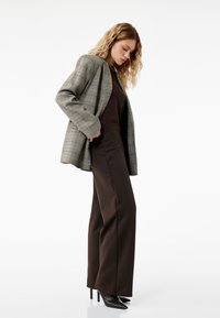 Rute oversize blazer i grå, parret med brune, brede bukser. Modellen bærer sorte spidse hæle og en tætsiddende mørk top.