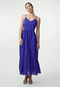 Vestido maxi roxo com alças finas, design de renda floral, barra com volantes e tecido texturizado. Usado com sapatos pretos e brancos estampados.