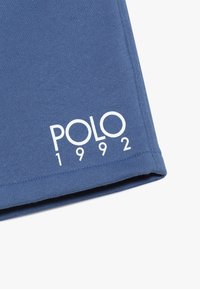Blå bomullstyg med en mjuk textur. Har vit tryckt text "POLO 1992" nära fållen. Klassisk t-shirt form med korta ärmar.