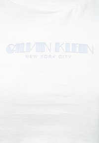 Fehér pamut póló dombornyomott világoskék "CALVIN KLEIN" logóval és "NEW YORK CITY" felirattal, kerek nyakkivágással és rövid ujjakkal.