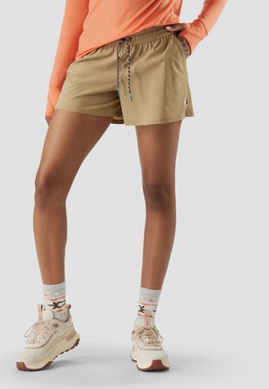 Persoon die beige korte broek met trekkoord, oranje trui met lange mouwen, patroon sokken en beige wandelschoenen draagt, staand tegen een grijze achtergrond.
