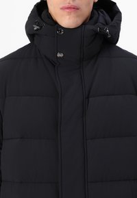 Veste noire matelassée avec un col haut, dotée de boutons-pression à l'avant et d'une capuche ajustable. Tissu texturé avec des détails en motif cousu.
