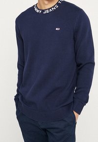 Laivastonsininen neulepaita, jossa on ribbikuviointi, pyöreä kaula-aukko, jossa on logolla varustettu nauha. Pienikokoinen logopatch vasemmassa rinnassa.