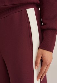 Bordó szűk szabású melegítőnadrág, fehér oldalsávval és textúrált anyaggal. Az egyik kéz a nadrág szárán pihen.