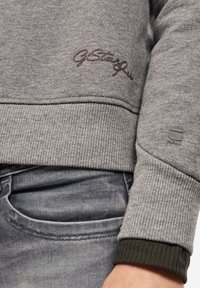 Sudadera gris con puños y dobladillo acanalados, con un logo bordado en marrón oscuro. Llevada sobre unos vaqueros azules, mostrando una tela texturizada.