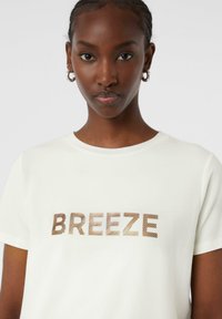 Weißes Baumwoll-T-Shirt mit einem strukturierten, bronzefarbenen "BREEZE"-Grafik auf der Vorderseite. Rundhalsausschnitt und kurze Ärmel, glatte Textur.