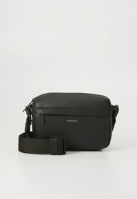 Borsa a tracolla in pelle nera testurizzata con tracolla regolabile e logo Calvin Klein frontale, su sfondo bianco semplice.