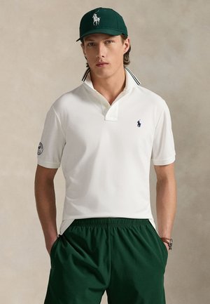 Ung mand iført en hvid polotrøje med oprejst krave, mørkegrønne shorts og en mørkegrøn kasket, stående med hænderne i lommerne.