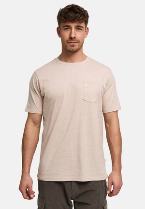 Mann trägt beigefarbenes T-Shirt mit kurzen Ärmeln, Brusttasche und kleinem Logo, kombiniert mit dunkler Cargohose, steht vor hellem Hintergrund.
