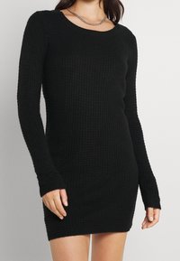 Abito lungo nero a maniche lunghe realizzato in tessuto a maglia testurizzata, con scollatura rotonda e silhouette aderente, che si estende fino a metà coscia.