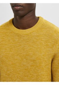 Gelber Strickpullover mit strukturiertem Muster und geripptem Rundhalsausschnitt. Der Stoff wirkt weich und warm, mit einer körpernahen Passform.