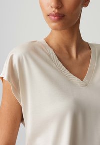 Lichtbeige top met V-hals en korte mouwen, gemaakt van een gladde stof. Kenmerkt zich door een geribbelde halslijn en een relaxed fit, die subtiele plooien accentueert.