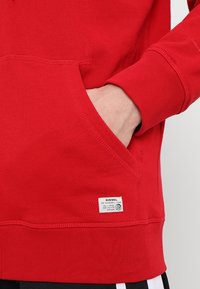 Main avec le poignet partiellement à l'intérieur de la poche avant d'un sweat-shirt rouge Diesel avec une étiquette de marque blanche près de l'ourlet inférieur.