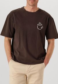 T-shirt marrone a maniche corte in cotone, con una semplice grafica bianca sul petto che rappresenta una tazza con fumi. Vestibilità rilassata, texture morbida.