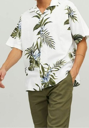 Mann trägt weißes kurzärmliges Hemd mit grünem tropischem Blattmuster und olivgrüne Hose, Hand in der Tasche, vor einfarbigem Hintergrund.