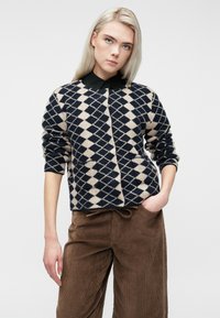 Jovem mulher com cabelo loiro longo, vestindo uma camisa preta com colarinho sob um pulôver em padrão de diamante azul-marinho e bege, e calças de courduroy castanhas.