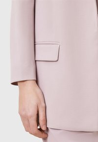 Blazer rosa chiaro visto di lato, con una singola tasca sul petto, texture liscia e vestibilità su misura con maniche leggermente arrotolate.