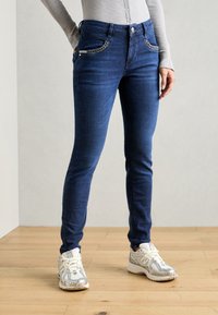 Uže pantalone u tamnoplavom denimu, s ukrašenim džepovima i kopčanjem na dugme. Uparene s svijetloplavim sportskim tenisicama.
