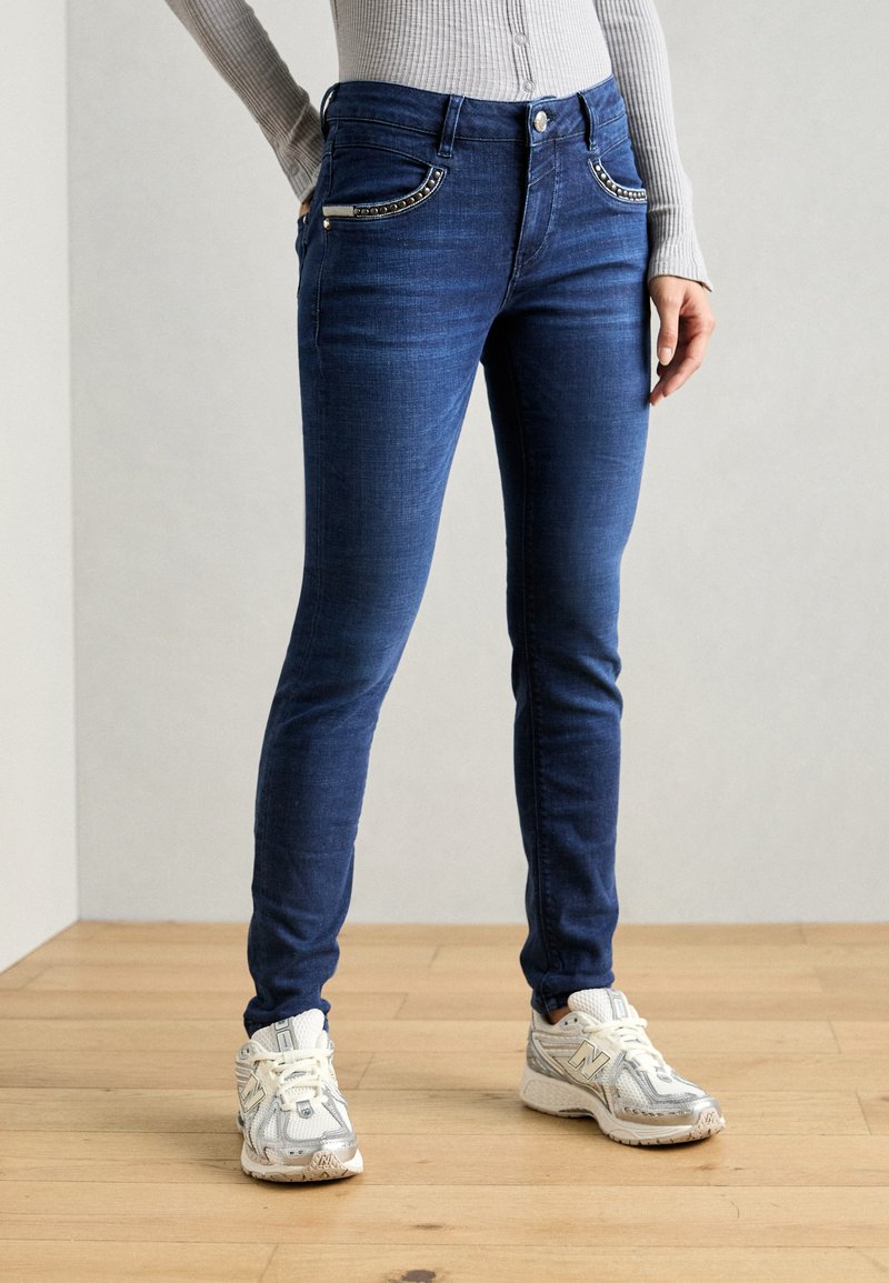 Uže pantalone u tamnoplavom denimu, s ukrašenim džepovima i kopčanjem na dugme. Uparene s svijetloplavim sportskim tenisicama.