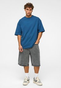 Blauwe oversized katoenen t-shirt en grijze denimshorts. Eenvoudig ontwerp, relaxte pasvorm met zichtbare stiksels. Witte sokken en met patroon versierde sneakers.