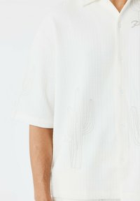 Camisa blanca de manga corta hecha de tela texturizada, con un sutil bordado de cactus en beige. Los botones son blancos y tienen un diseño de cuello relajado.