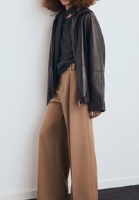 Giacca di pelle marrone sopra un top grigio scuro e pantaloni beige a gamba larga. L'outfit presenta una vestibilità ampia e dettagli di texture sottili.